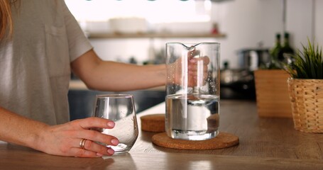 Unrecognizable young girl or woman holds glass jar water in kitchen. Hand of anonymous person in white shirt take jug from countertop