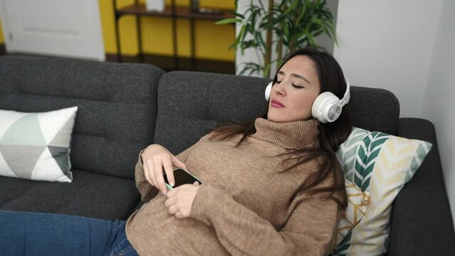 Beautiful hispanic woman listening to music with smartphone lying on sofa at home