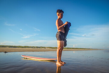 Young preteen surfer standing on beach putting on wetsuit with surfboard at his feet
