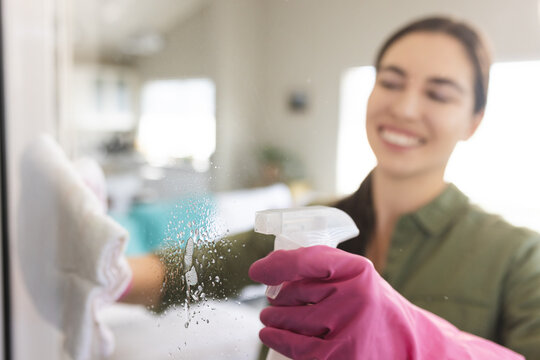 Caucasian young woman spraying rubbing alcohol on glass wall and wiping with towel at home