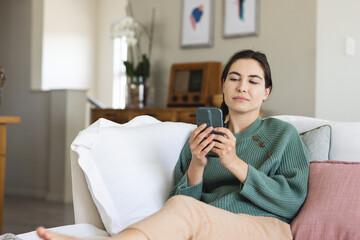Caucasian young woman using social media over smart phone while relaxing on sofa at home