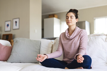 Caucasian young woman with eyes closed meditating while sitting on sofa in living room, copy space