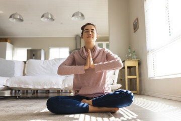 Caucasian young woman meditating in prayer position while sitting on floor in living room