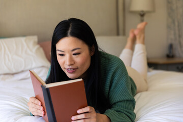 Happy asian woman lying on her bed reading a book during the day