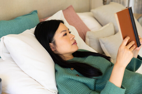 Happy Asian Woman Lying On Bed With Head On Pillows, Reading A Book During The Day