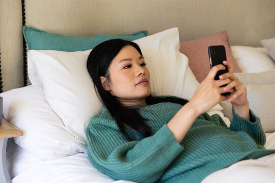 Thoughtful asian woman lying on bed with head on pillows using smartphone