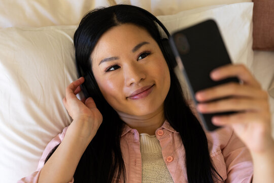 Smiling asian woman lying on bed wearing headphones using smartphone - Powered by Adobe