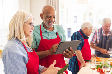 Happy group of diverse senior friends in aprons, using tablet during cookery class