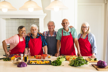 Portrait of happy, diverse senior friends wearing aprons in kitchen, embracing during cookery class