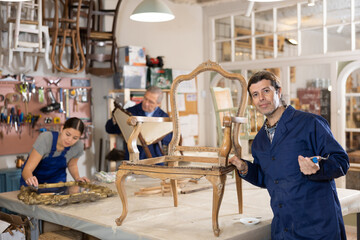 Professional middle-aged craftsman restorer in uniform renewing antique chair while working in...