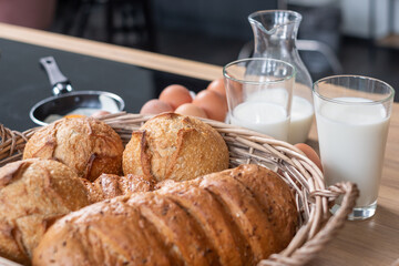Fresh loaves of bread in the basket on the table. White milk in a transparent cup.  The eggs on the table by the stove. A freshly fried egg in the pan on the stove.