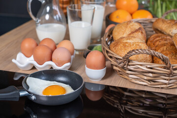 Various breads in baskets, 2 glasses and a mug with milk, oranges in a black dish and some eggs on the wooden table. Bull's eye on the pan