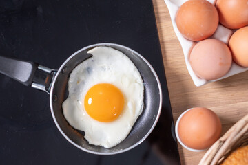 The freshly fried egg in a tiny black pan on the stove.  The fresh eggs in white dishes, milk, some bread in a basket in the background. Copyspace