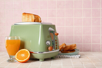 Bread slices in modern toaster, cup of juice, orange and toasts on tile table