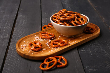 Board with bowl of tasty salted pretzels on dark wooden background