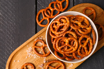 Board with bowl of tasty salted pretzels on dark wooden background, closeup - obrazy, fototapety, plakaty