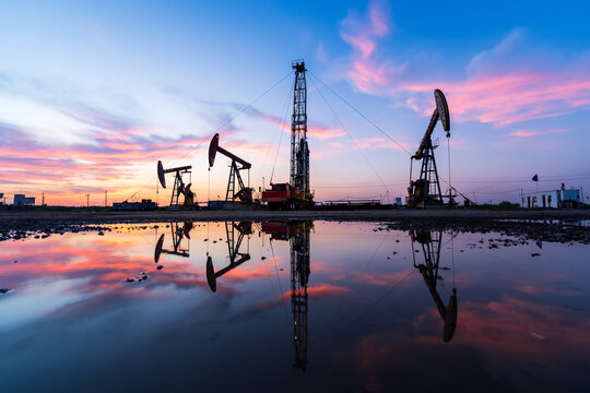 in the evening, oil pumps are running, The oil pump and the beautiful sunset reflected in the water, the silhouette of the beam pumping unit in the evening. - Powered by Adobe