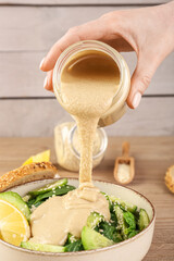 Woman adding tahini into bowl of salad on wooden table, closeup