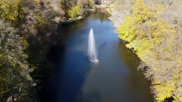 Landscaped park with lake, fountain, trees, large stones and lot of people walking on sunny autumn day. Trees with yellow leaves on shores, tourists, high flowing jet of fountain. Aerial drone view