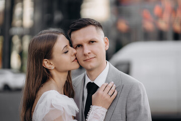Front view of bride and groom getting married, having photoshoot outdoors. Woman wearing wedding dress, kissing groom, man looking at camera. Concept of marriage and love.