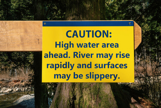 Trail caution sign about high water and fast rising river. Yellow large sign on fence pole in front of river from the park authority. Trail and hiking safety sign. Selective focus.