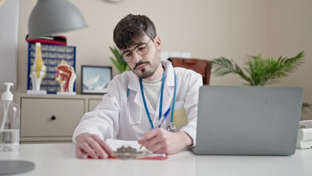 Young hispanic man doctor using laptop writing on clipboard at clinic