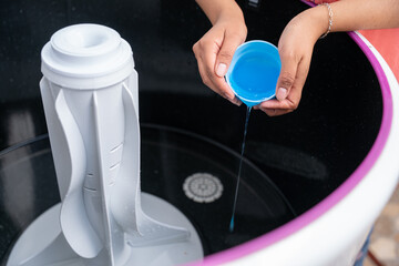 A young woman is pouring some liquid detergent into a top load washing machine