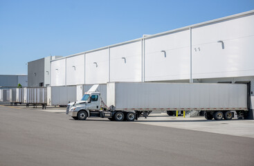Bonnet day cab white big rig semi truck with roof spoiler and dry van semi trailer loading cargo standing in warehouse dock gate at sunny working day