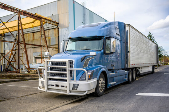 Blue Gray Big Rig Semi Truck With Aluminum Grille Guard And Refrigerator Semi Trailer With Open Back Door Standing Near The Warehouse On The Street At Industrial Area Waiting For The Next Load