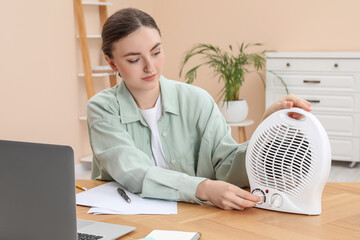 Young woman adjusting temperature on modern electric fan heater at wooden table indoors