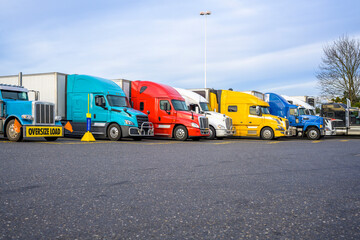 Fototapeta na wymiar Different big rigs semi trucks with semi trailers standing in row on the reserved spots on truck stop parking lot