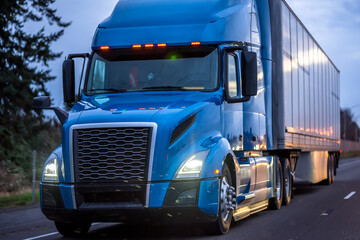 First drops of rain in the beams of the headlights of the blue big rig semi truck with dry van semi trailer driving on the evening twilight highway road