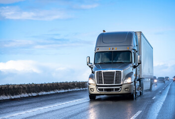 Gray big rig semi truck with turned on headlights transporting cargo in dry van semi trailer...