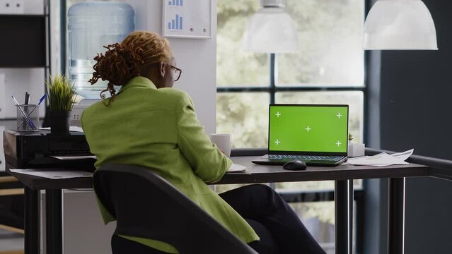 African american woman analyzing laptop with greenscreen, using isolated display at office desk. Company worker checking blank chroma key mockup template at executive business job.