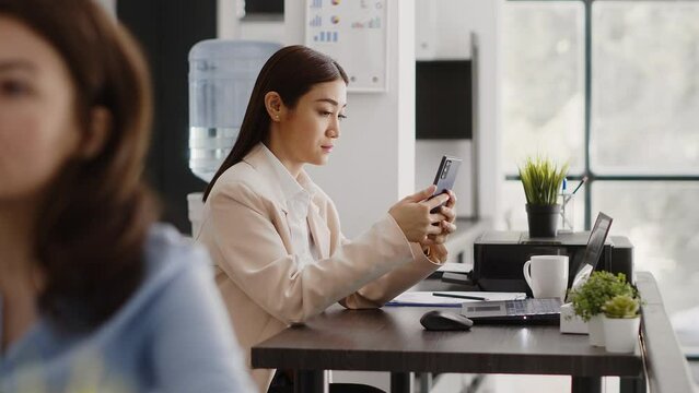 Asian office worker using smartphone app in workplace, planning online report before taking break at desk. Young employee browsing internet and social media app in coworking space.