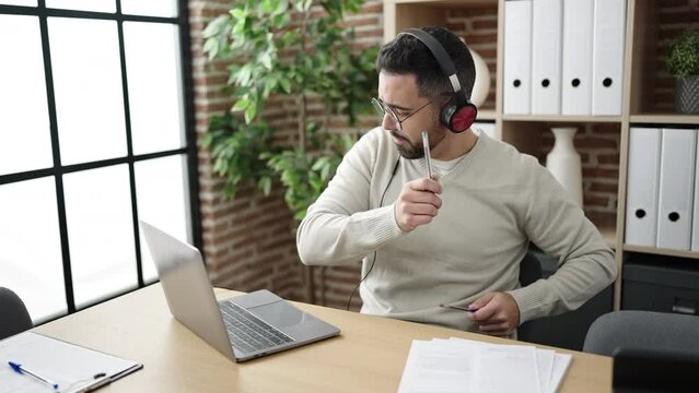 Young hispanic man business worker listening to music doing drummer gesture at office