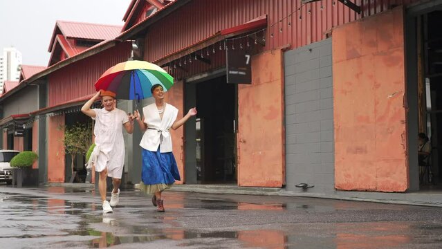 Asian drag queen gay friends in woman clothes holding umbrella walking down city street together in raining day. Diversity sexual equality, lgbtq pride people and transgender cross-dressing concept.