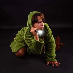 Young frightened preteen boy crouching on ground wearing pajamas and robe with flashlight and teddy...