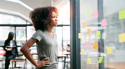 Pensive young businesswoman standing next to a glass wall, looking at sticky note.