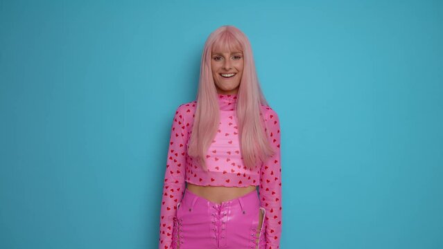 Beautiful model with pink hair and in pink clothes standing in the midle of blue background, shakes the head, shows thumb up and smiles happily showing enjoyment, copy space