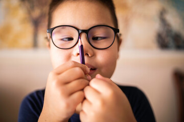Smart young Asian boy wearing glasses squints at the pencil. The vision diseases problem. - obrazy, fototapety, plakaty