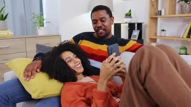 Black couple, phone and relax on sofa in home living room with comic smile, laugh and social media video. Man, woman and smartphone texting with love, bond and funny meme on web, app ux and internet