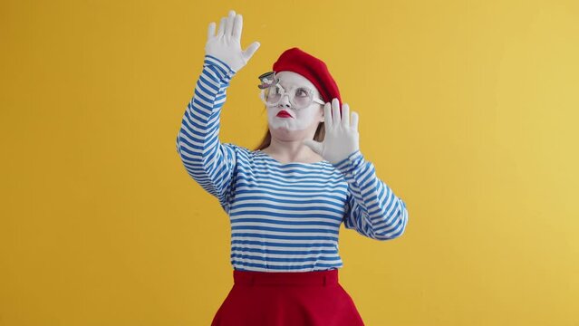 Female mime with perplexity emotion performs performance in an invisible box, front view, yellow background. Woman in suit and makeup mime pretends that she is behind an invisible wall