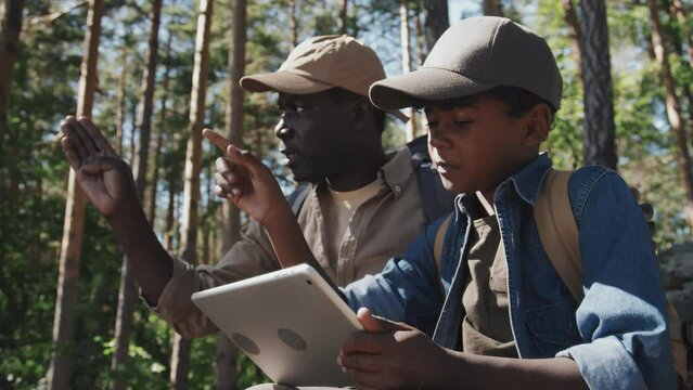 Low angle medium shot of African American boy spending weekend with his grandpa hiking in mountains choosing path with use of map app on digital tablet