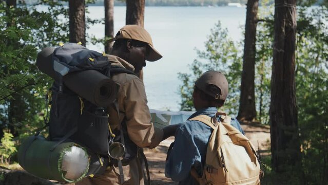 Rear view of African American grandpa and his grandson wearing backpacks hiking together in mountains using map to choose direction
