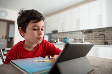 Adorable teenager, elementary school student on distance learning, sitting at desk, doing homework, studying online, watching webinar on digital tablet, holding a pen and making notes in his textbook