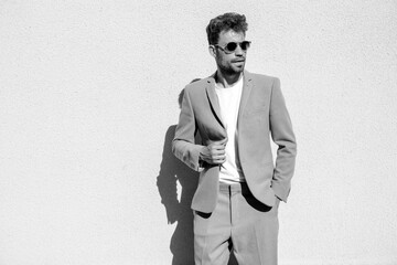 Handsome confident stylish hipster model.Sexy modern man dressed in elegant beige suit. Fashion male posing in the street background in Europe city at sunset. In sunglasses. Near white wall