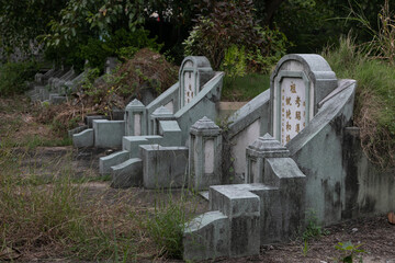 Old Chinese cemetery in Bangkok. A row of gray stone tombstones with Chinese hieroglyphs. Gloomy atmosphere and twilight lighting. Place is abandoned, fallen broken branches and leaves on the ground