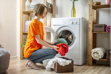 Woman is puting multicolor clothes into the washing machine in the laundry room. The concept of caring for things, cleaning and hygiene - 584156642