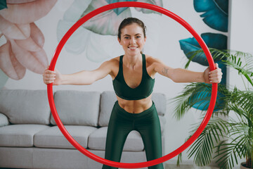 Young strong sporty athletic fitness trainer instructor woman wear green tracksuit hold look through hula hoop for slim waist training do exercises at home gym indoor Workout sport motivation concept